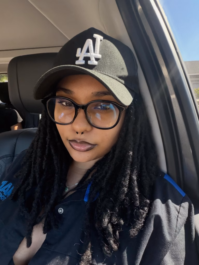 Locs Worn Down Under a Baseball Cap With Black Round Frames